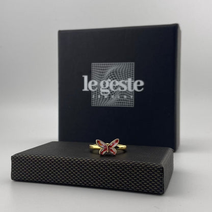 A gold ring sitting on a box. The face of the ring is a butterfly with pink and white marble wings.